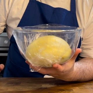 Un uomo con un grembiule tiene in mano una ciotola di pasta avvolta in pellicola trasparente