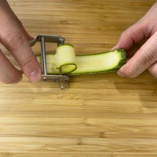 Una persona sta sbucciando un cetriolo con un pelapatate su un tagliere di legno.