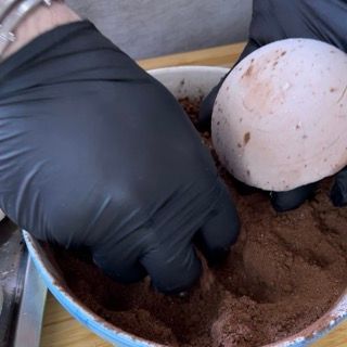 Una persona che indossa guanti neri tiene in mano un uovo in una ciotola di cioccolato in polvere