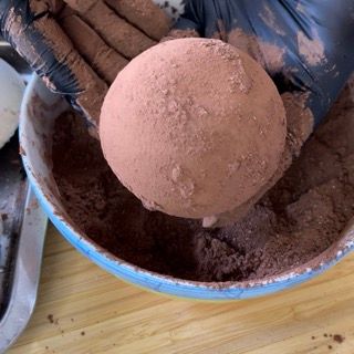 Una persona che indossa guanti neri tiene una palla di cacao in polvere sopra una ciotola di cacao in polvere