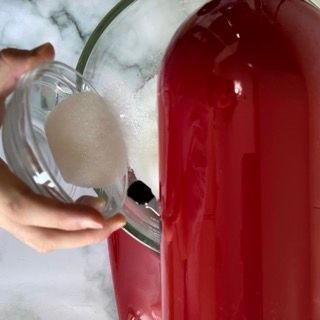 Una persona versa dello zucchero in una ciotola di vetro accanto a una bottiglia rossa.