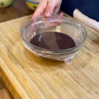 Una persona tiene in mano una ciotola di salsa al cioccolato su un tagliere di legno.