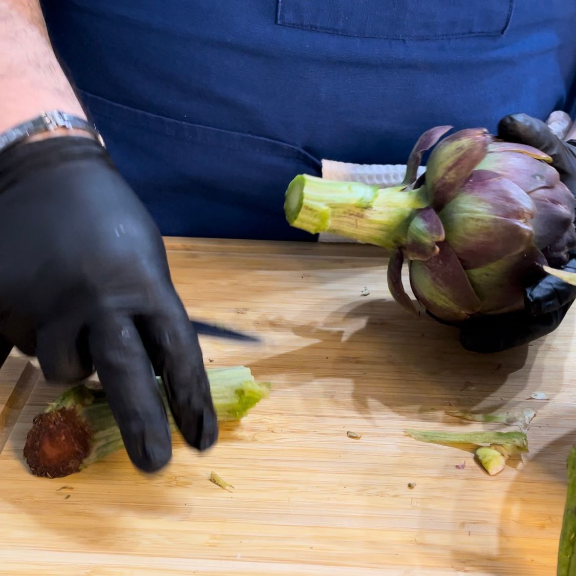 Una persona che indossa guanti neri sta tagliando un carciofo su un tagliere di legno.