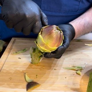 Una persona che indossa guanti neri sta tagliando un carciofo su un tagliere di legno.