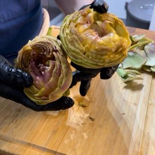 Una persona che indossa guanti neri tiene due carciofi su un tagliere.