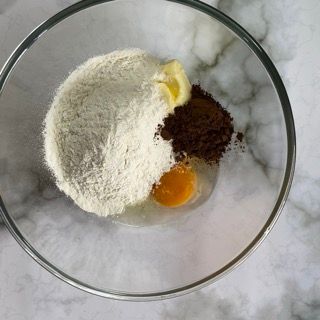 Una ciotola di vetro piena di farina, cacao in polvere, burro e un uovo.