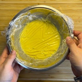 Una persona tiene in mano una ciotola piena di liquido giallo avvolto in pellicola trasparente.