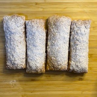 Quattro rotoli di pasta sfoglia ricoperti di zucchero a velo sono appoggiati su un tagliere di legno.