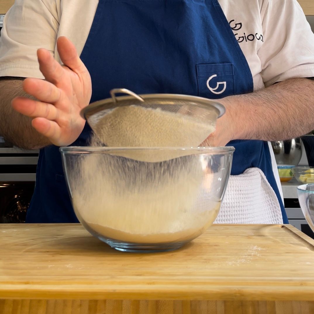 Un uomo con un grembiule blu sta setacciando la farina in una ciotola di vetro