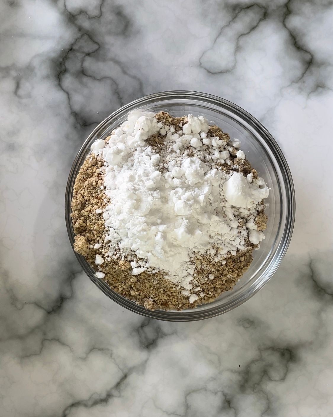 Una ciotola di vetro piena di zucchero a velo e semi di sesamo su una superficie di marmo.