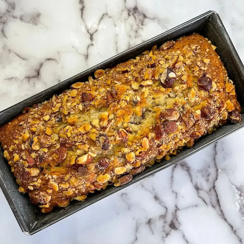 Banana bread soffice e umido appena sfornato, con gocce di cioccolato e frutta secca visibili in superficie.