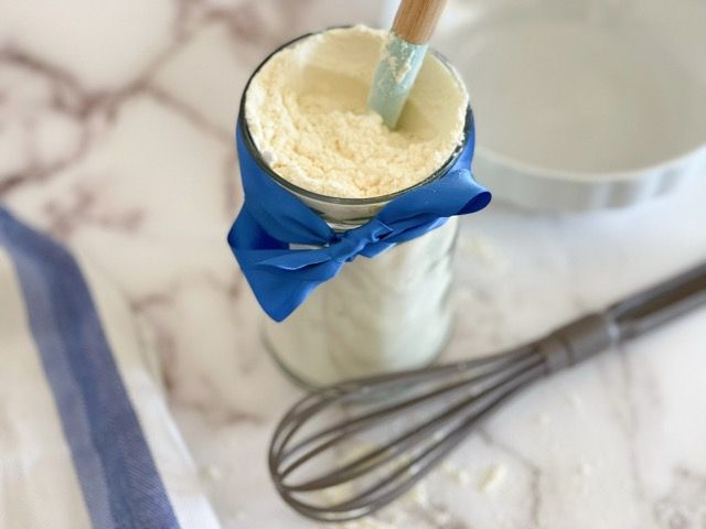 Un barattolo di farina con un fiocco blu e una frusta su un tavolo.