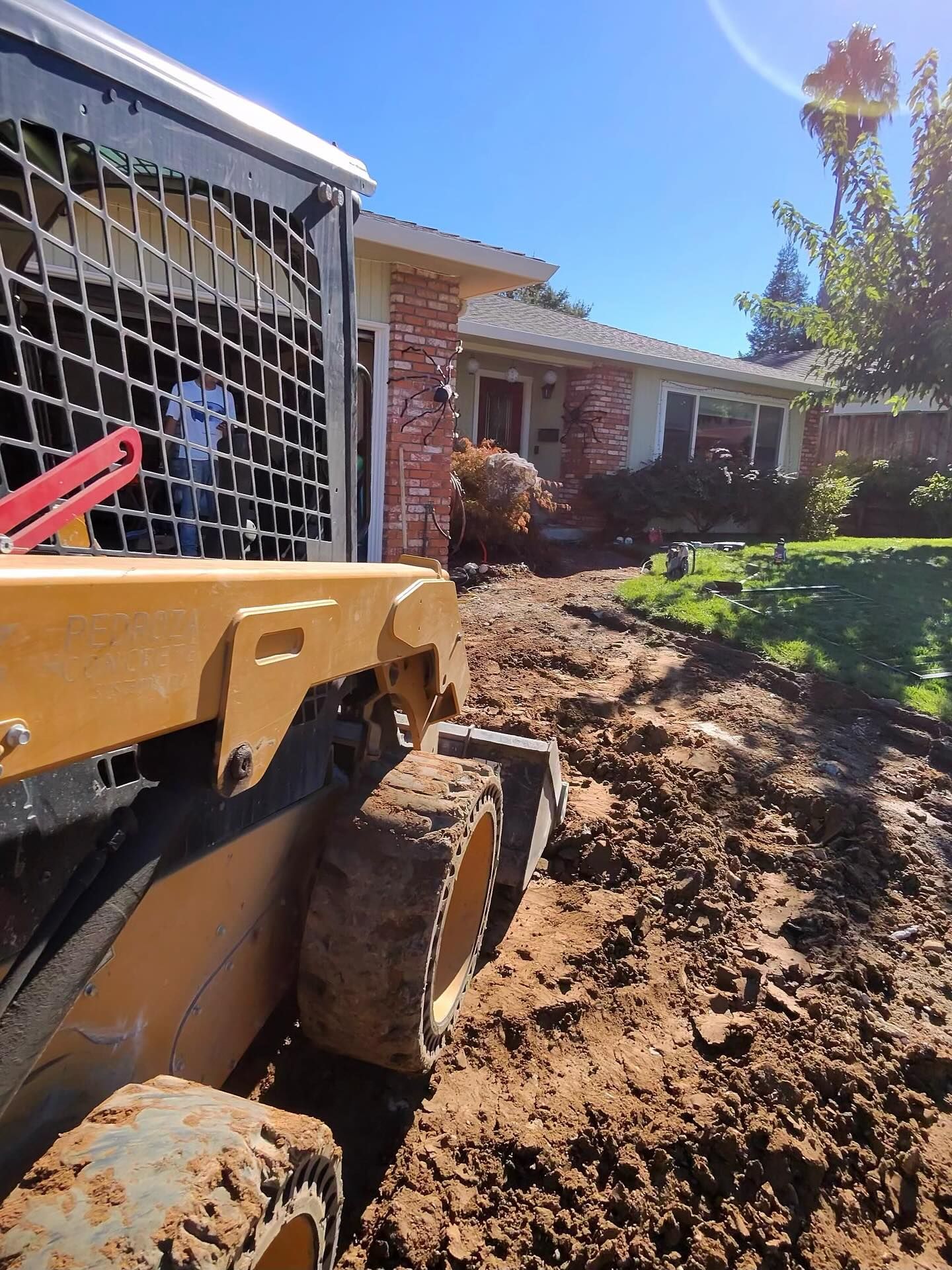 A yellow skid steer on a dirt yard in front of a brick-accented house on a sunny day.