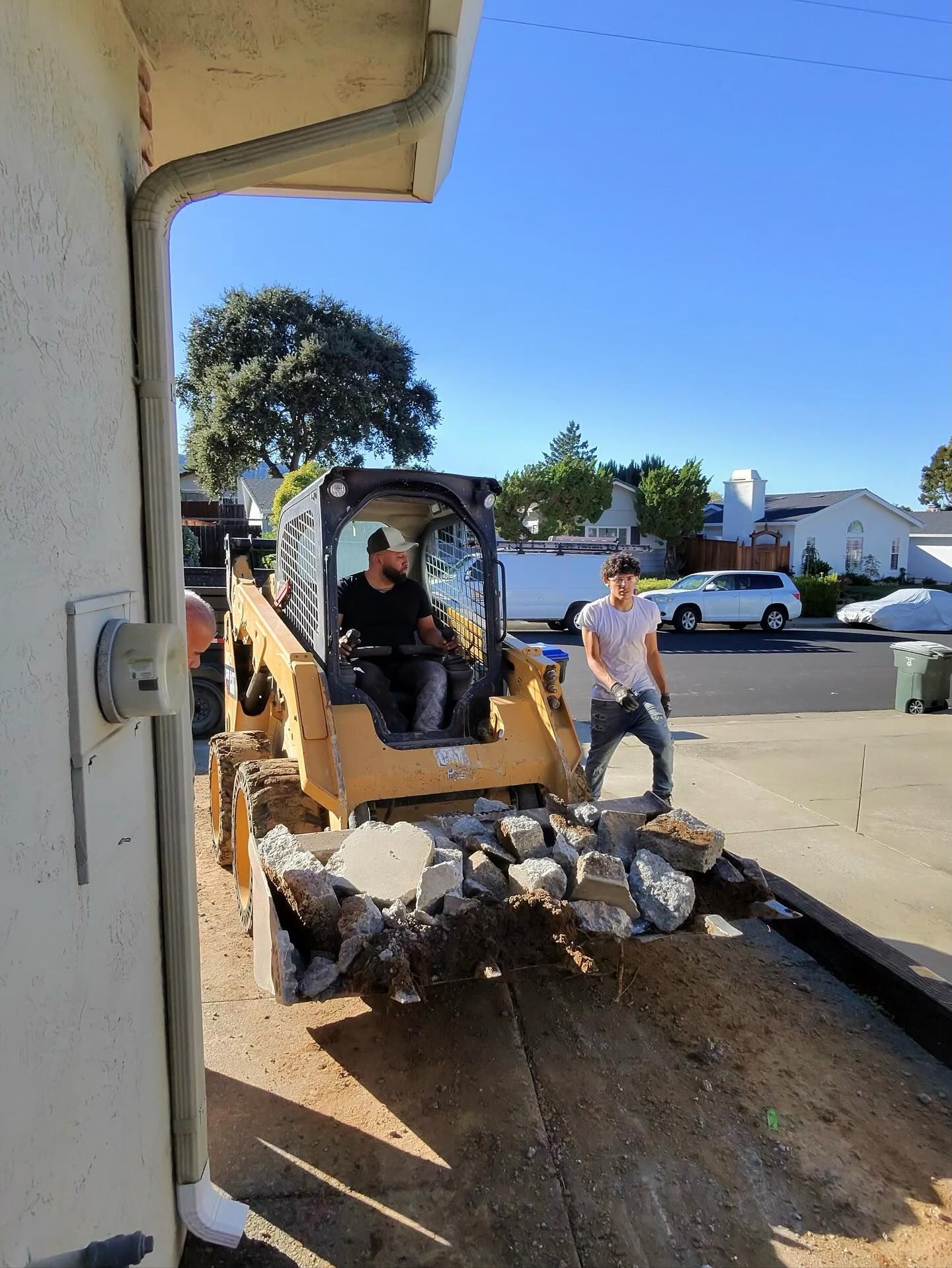 Two workers use a yellow skid steer to clear chunks of broken concrete from a residential driveway on a sunny day.