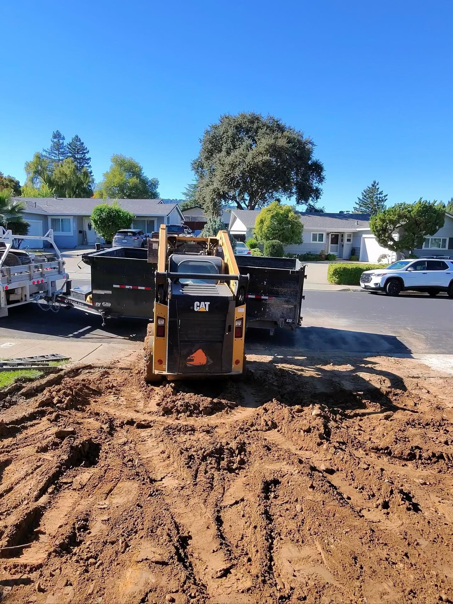 A yellow CAT skid-steer loader sits on a muddy, excavated residential yard, facing a large black dumpster in a driveway.