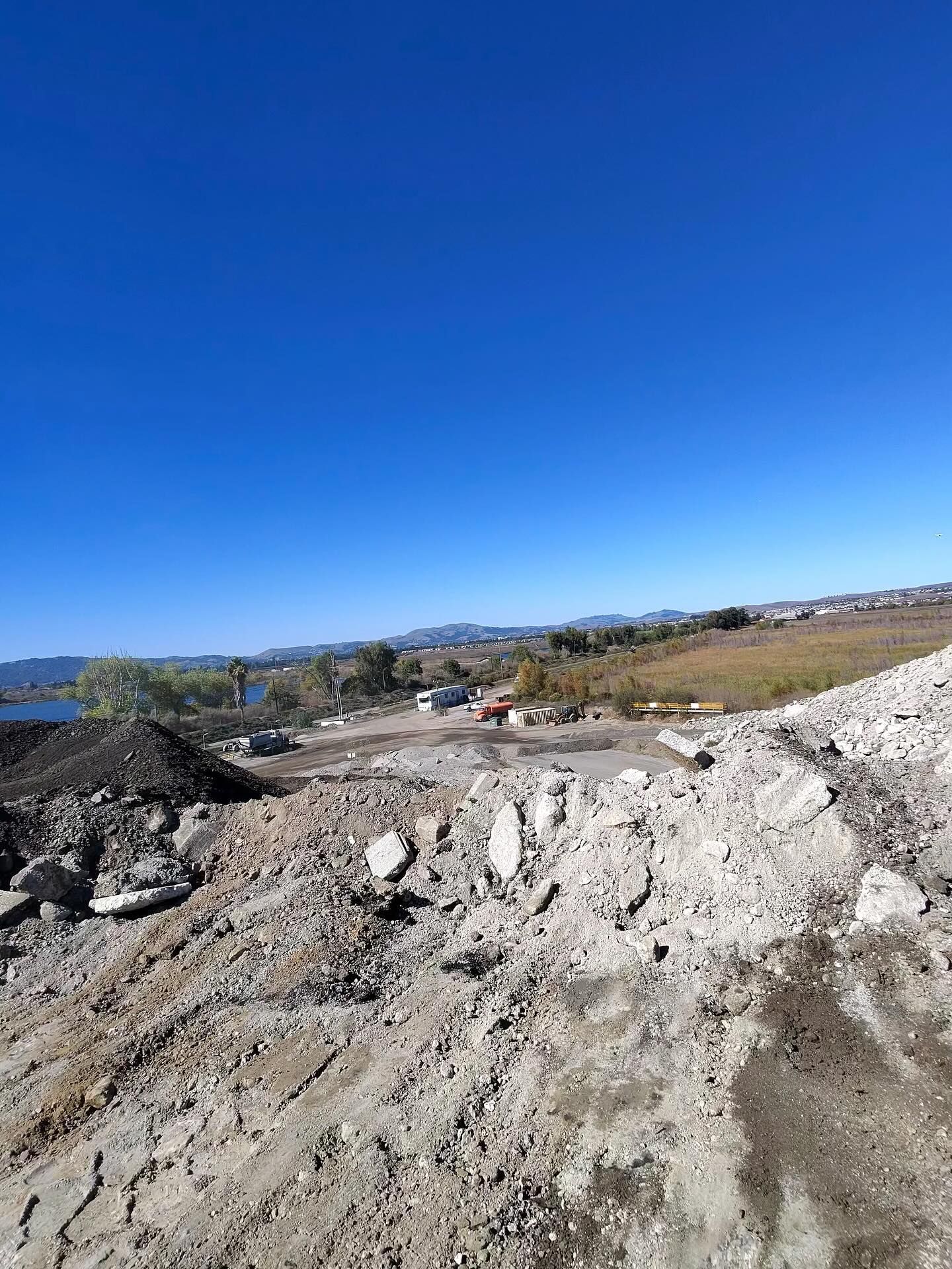 A dirt and concrete construction site under a clear blue sky, with a river and distant hills in the background.