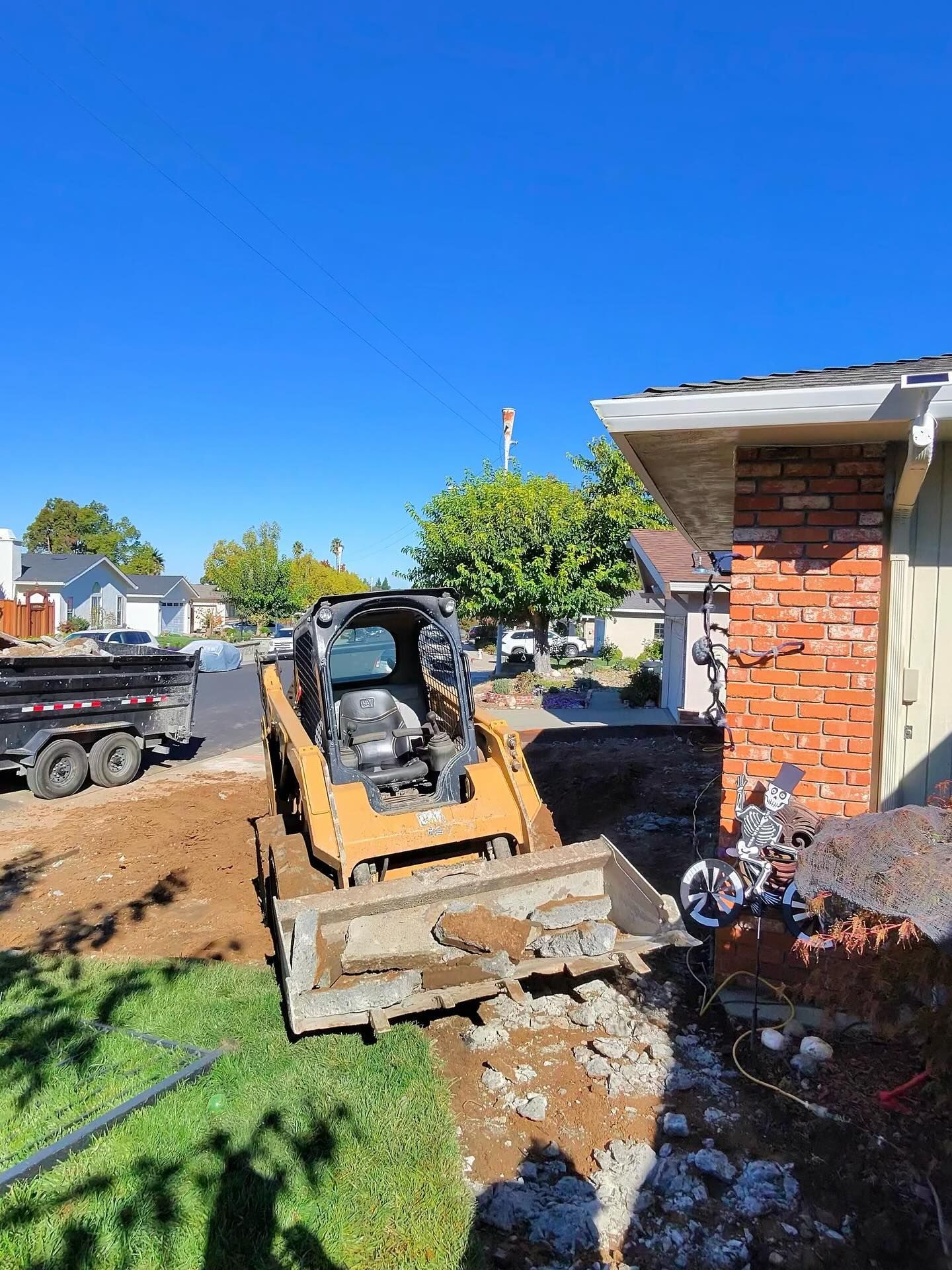 A yellow skid steer loader sits on dirt in a residential yard next to a brick house and a utility trailer.