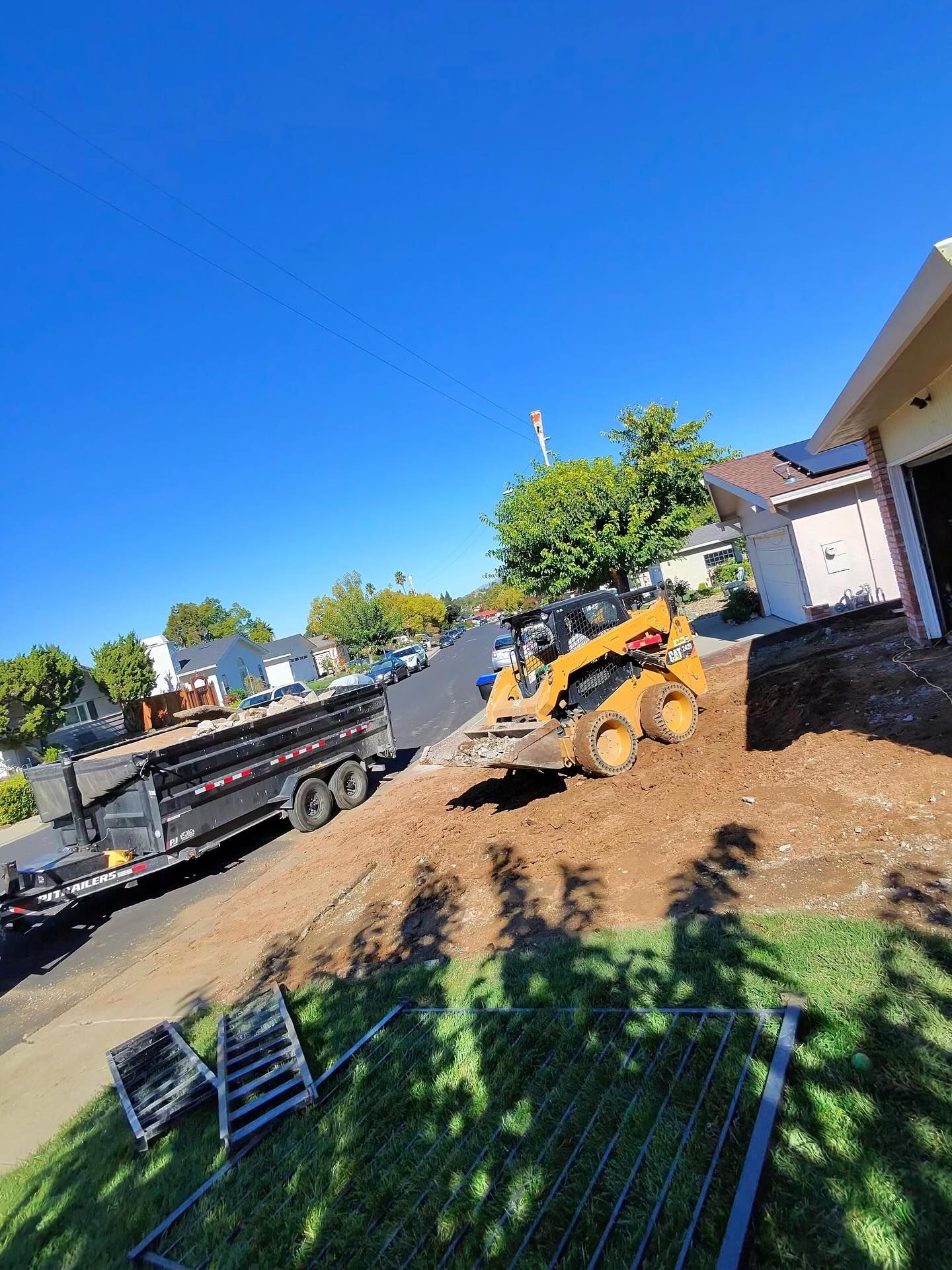 A yellow skid-steer loader works in a residential front yard near a parked dump trailer on a sunny day.