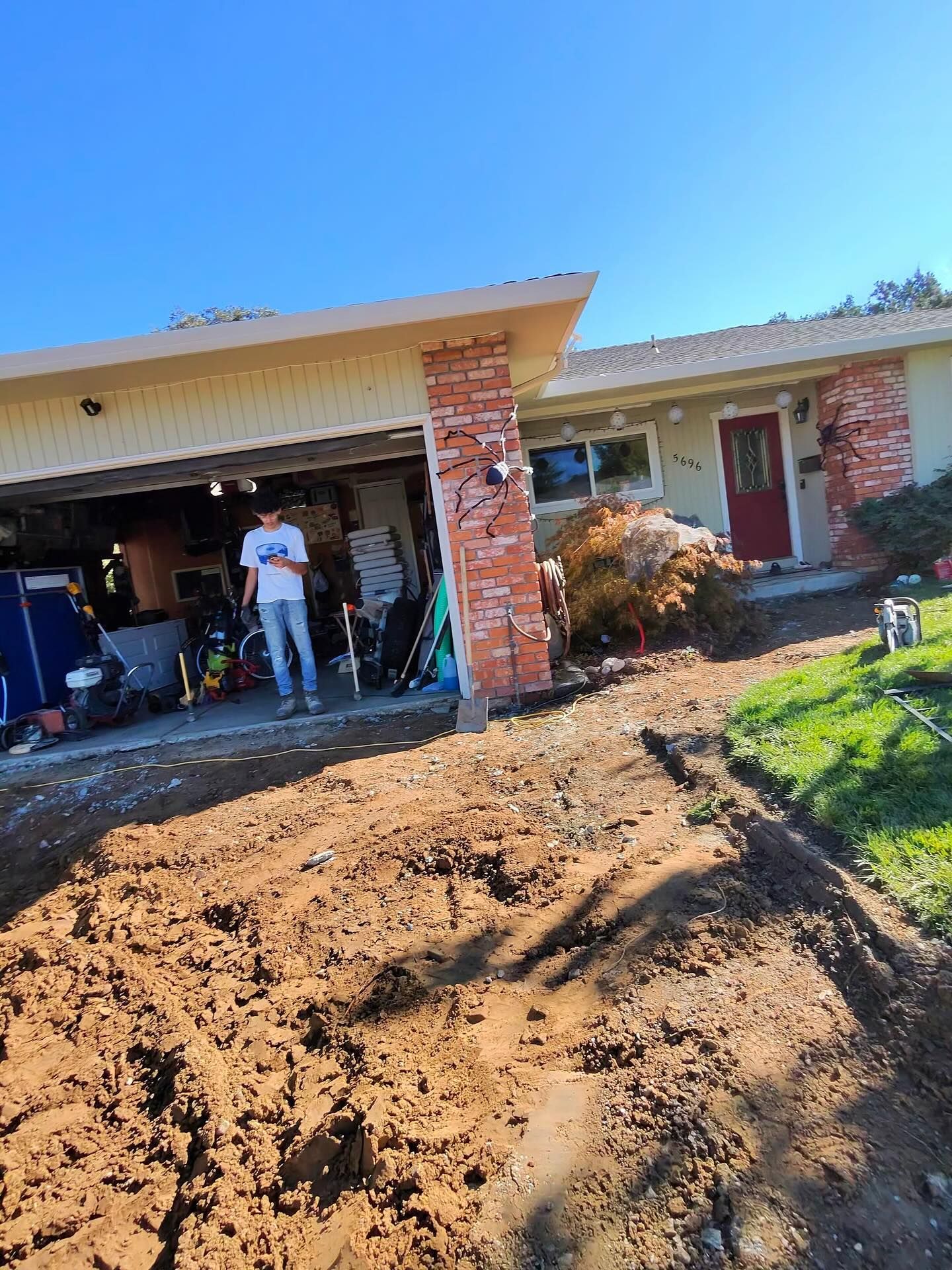 A person stands in a garage beside an unpaved, dirt-covered driveway in front of a suburban house with a red front door.