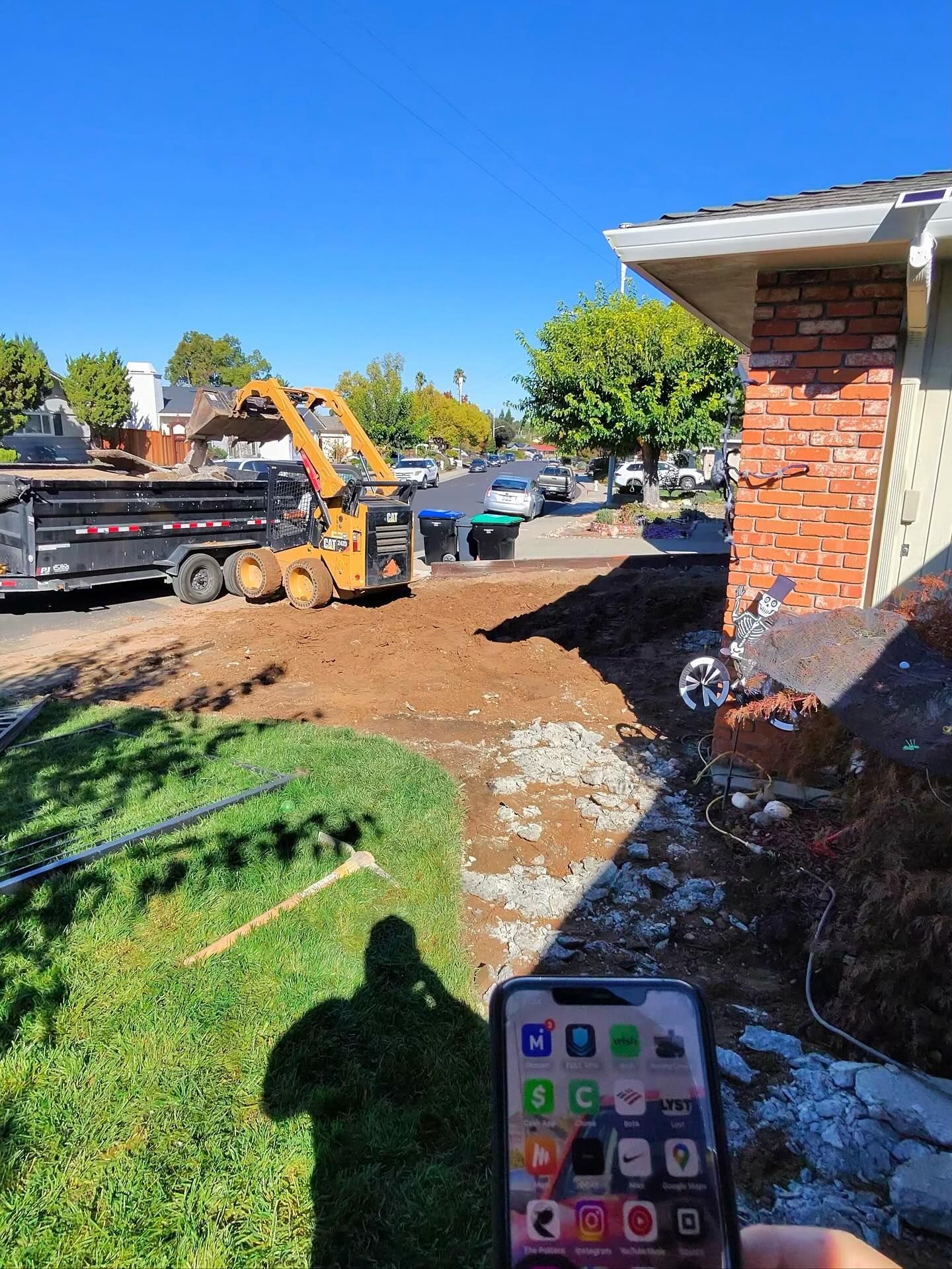 A yellow skid-steer loader loads dirt into a dump truck in front of a brick house, captured by a person holding a phone.