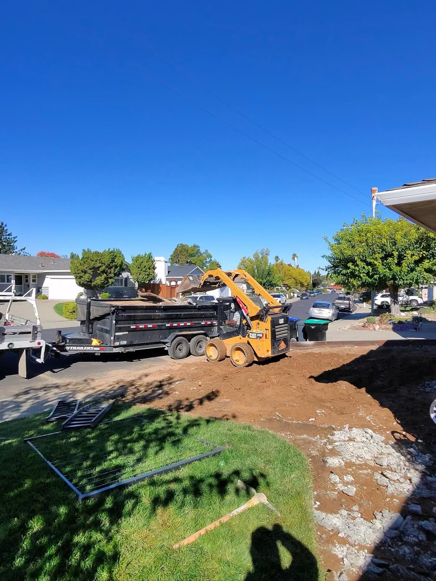 A yellow skid-steer loader fills a black dump trailer with dirt in a suburban front yard on a sunny day.