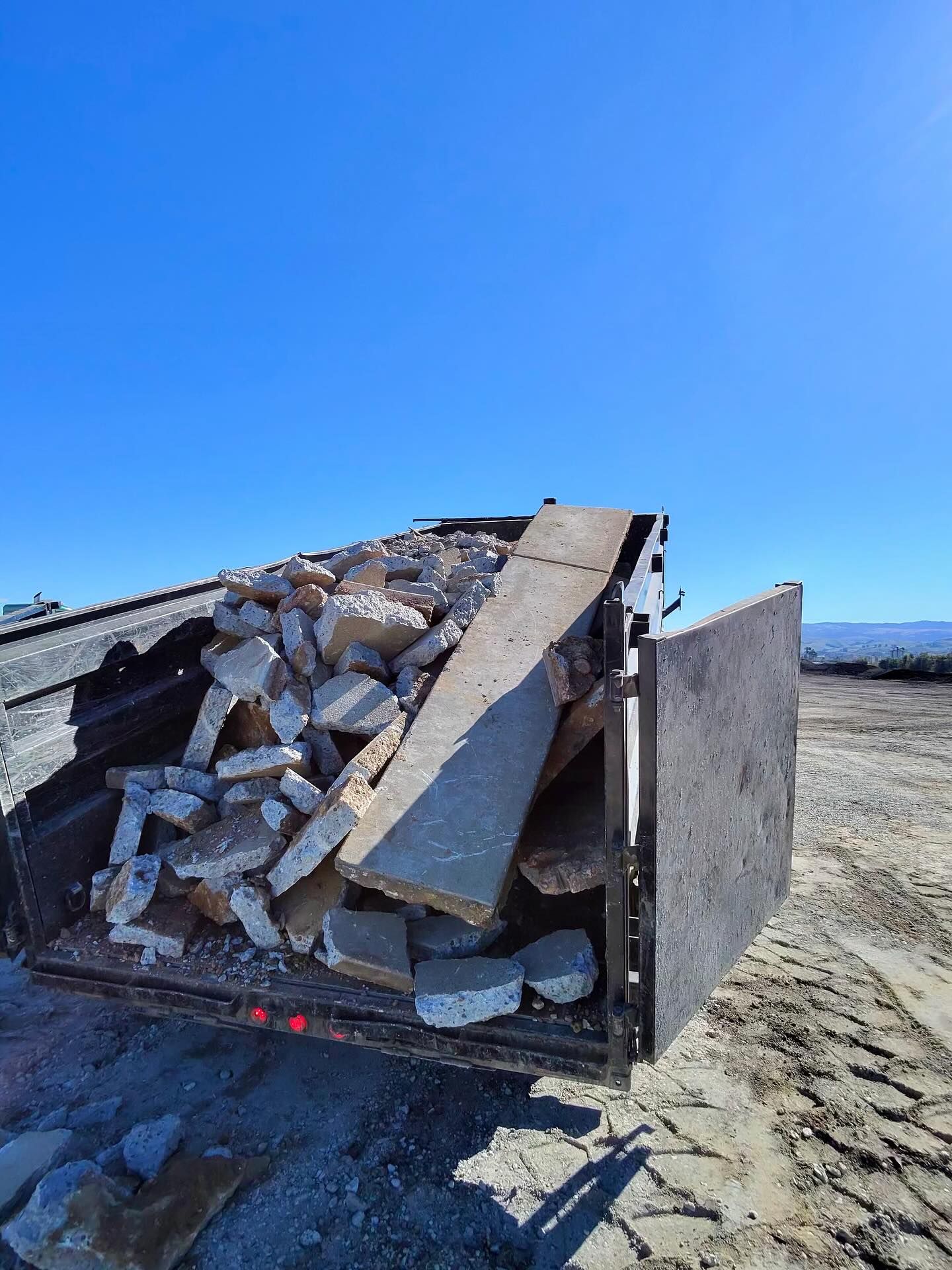 A metal dump trailer filled with broken concrete rubble sits on a dirt lot under a clear blue sky.