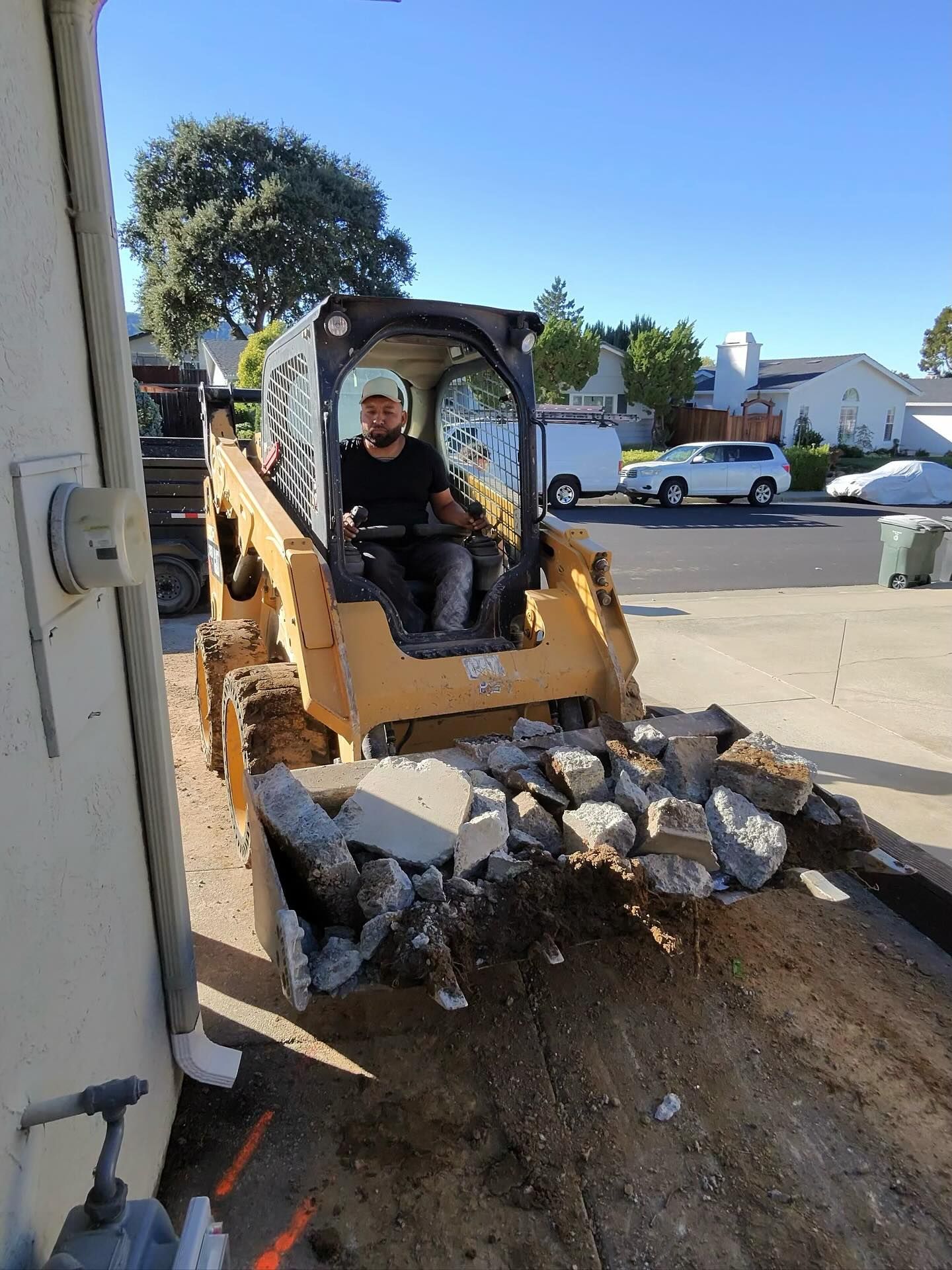 A person operates a yellow skid-steer loader carrying broken concrete debris in its bucket in a sunny suburban street.