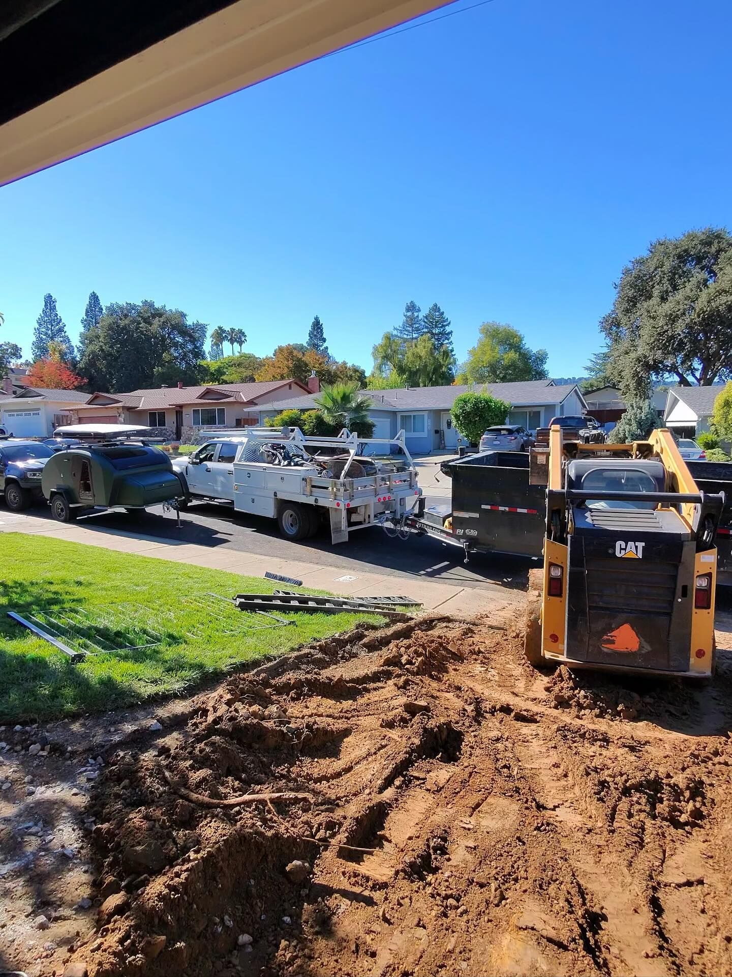 A yellow skid-steer loader sits next to a pile of dirt in a residential street, with service trucks parked nearby.