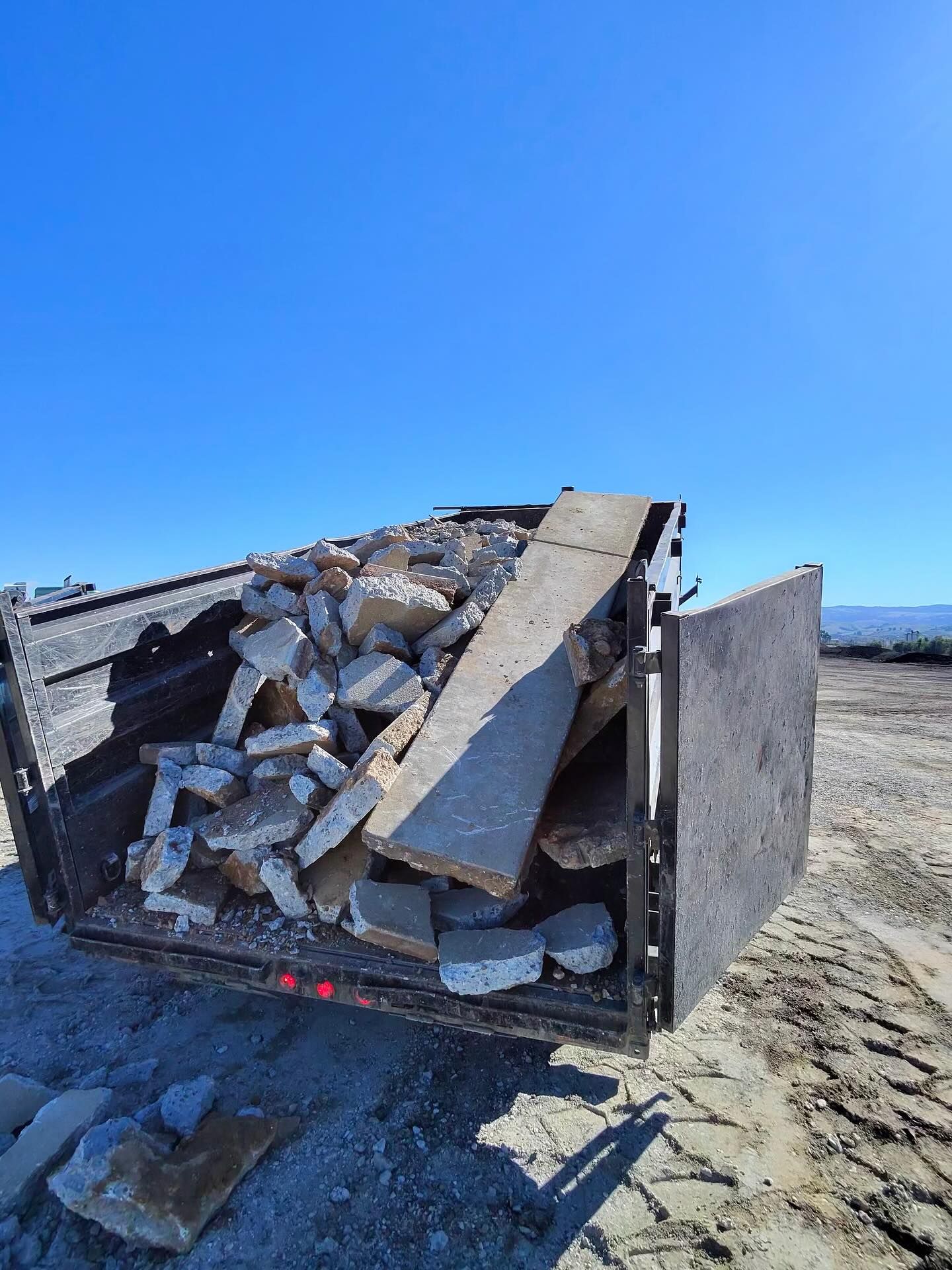 A trailer filled with broken concrete debris, chunks of rubble, and a long wooden plank under a clear blue sky.