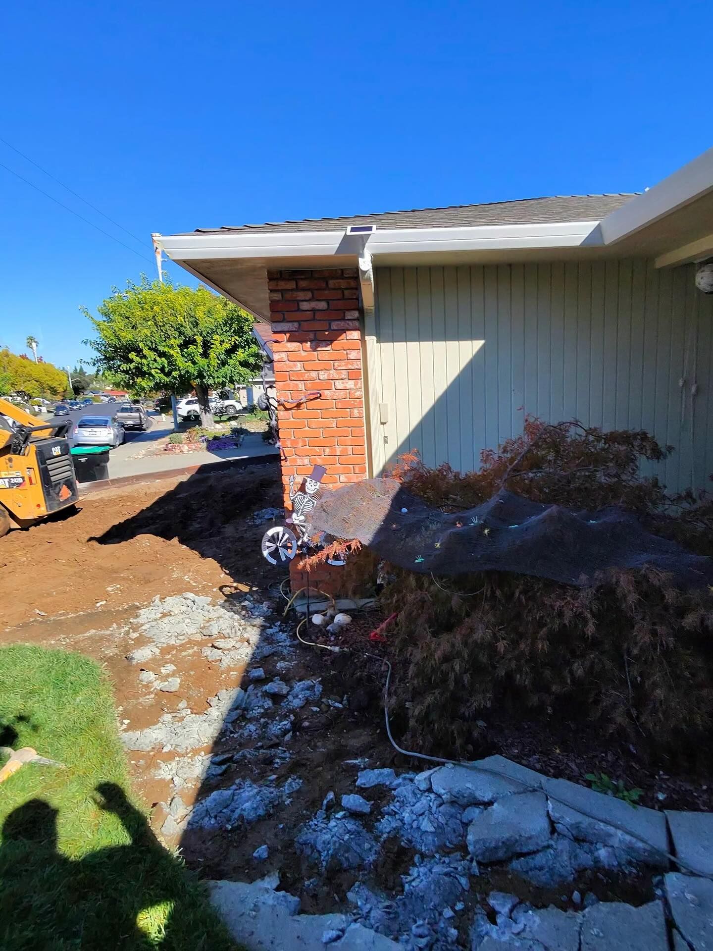 A front yard under construction with a yellow skid steer, piles of dirt, and rubble near a house with a brick corner.