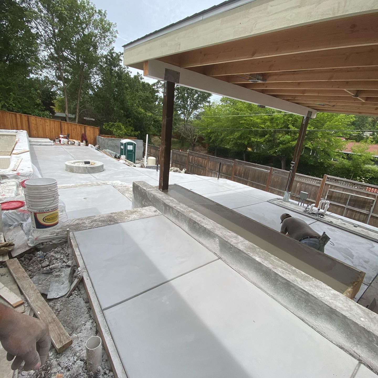 A construction site shows fresh concrete pathways and a patio cover under a wooden roof, with a worker smoothing cement.