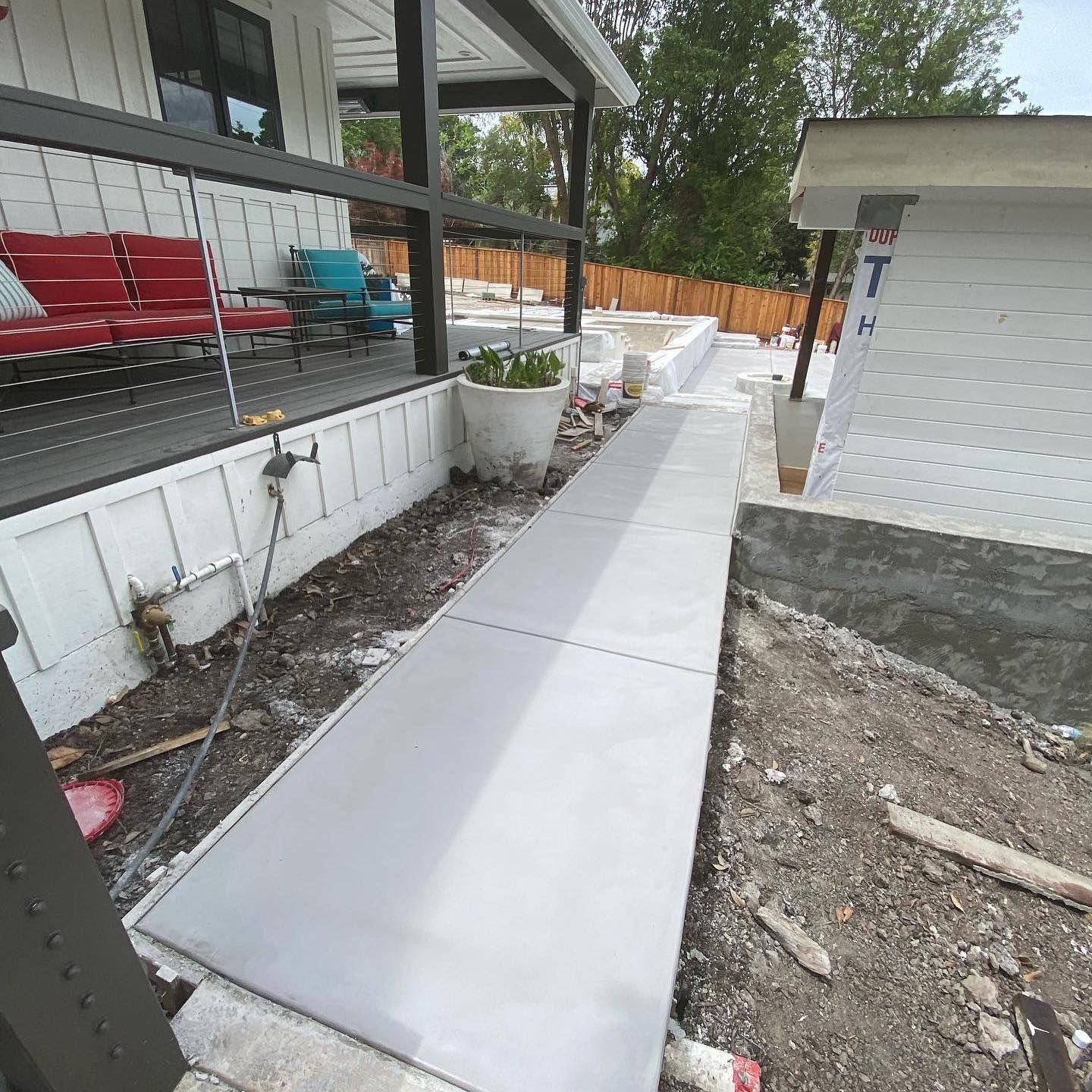 A newly poured concrete walkway runs between a porch with red seating and a building under construction.