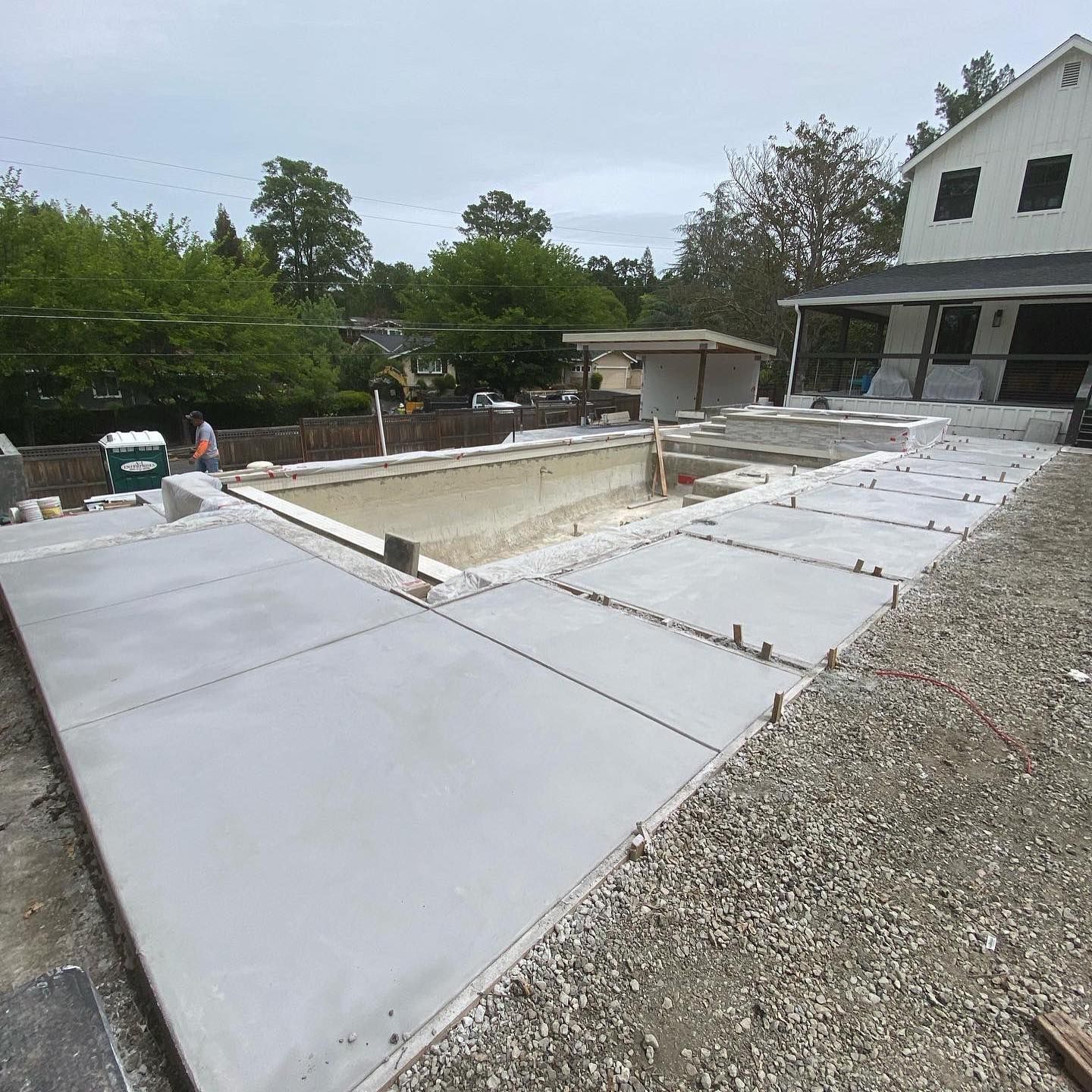 A backyard construction site featuring a partially installed swimming pool surrounded by newly poured concrete decking.