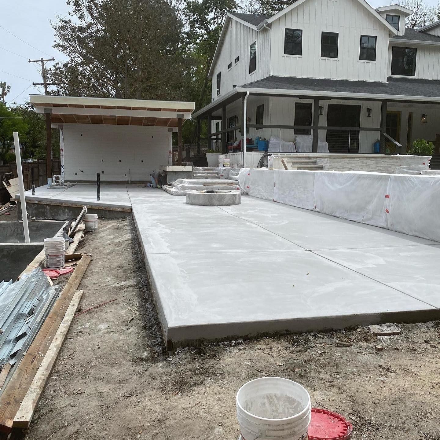 A backyard construction site features a newly poured concrete patio, a covered structure, and a white farmhouse style home.
