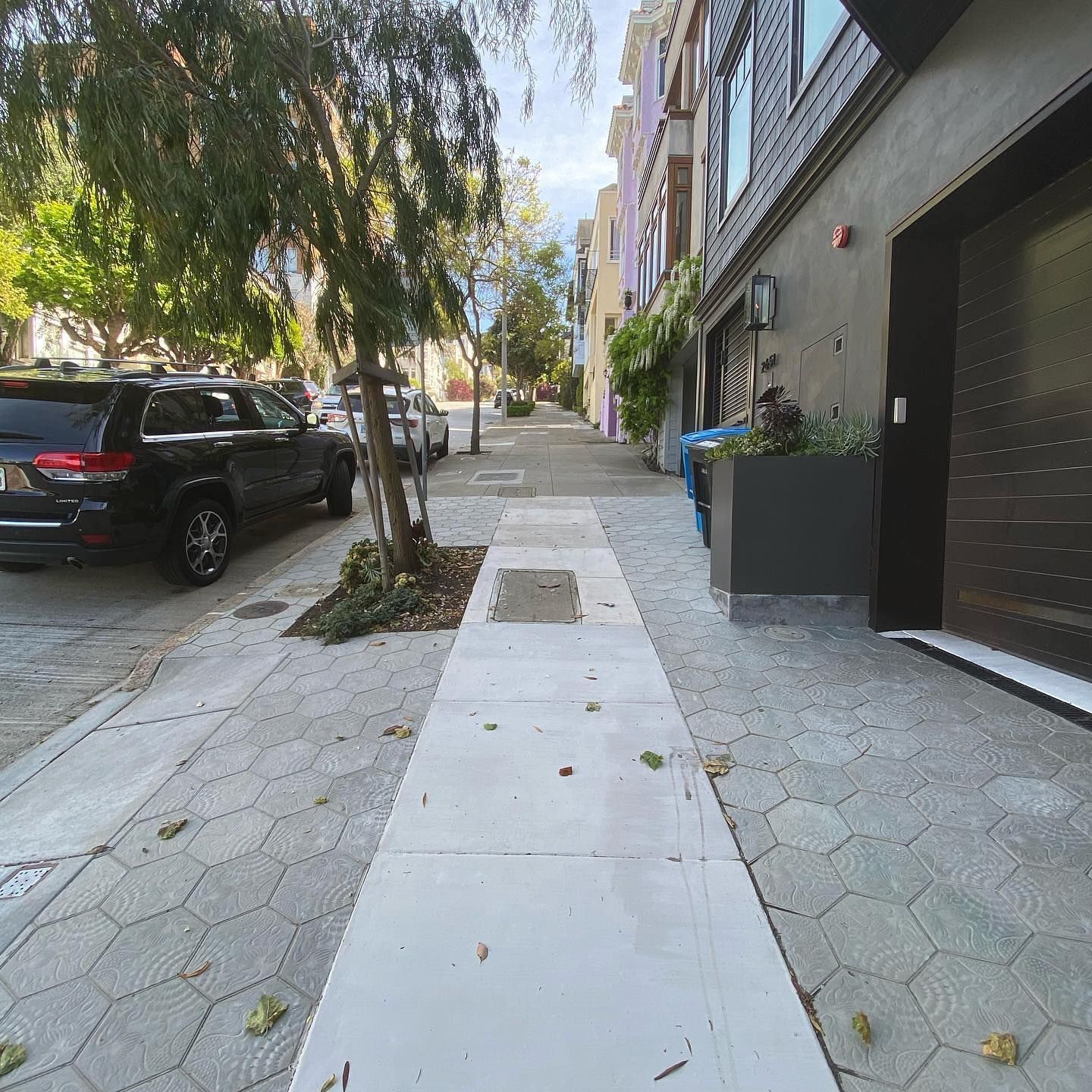 A sidewalk with a concrete path flanked by gray pavers, a black SUV, a tree, and a modern building entrance.
