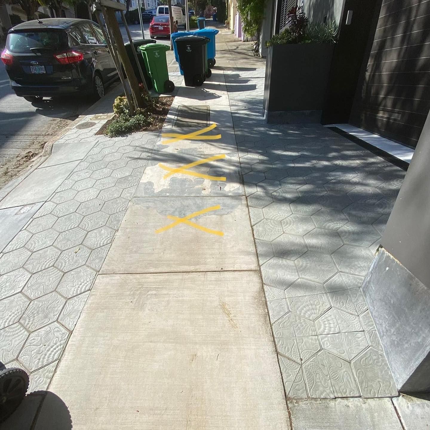 A sidewalk with several bright yellow 'X' marks painted on the concrete, leading toward a row of trash bins.