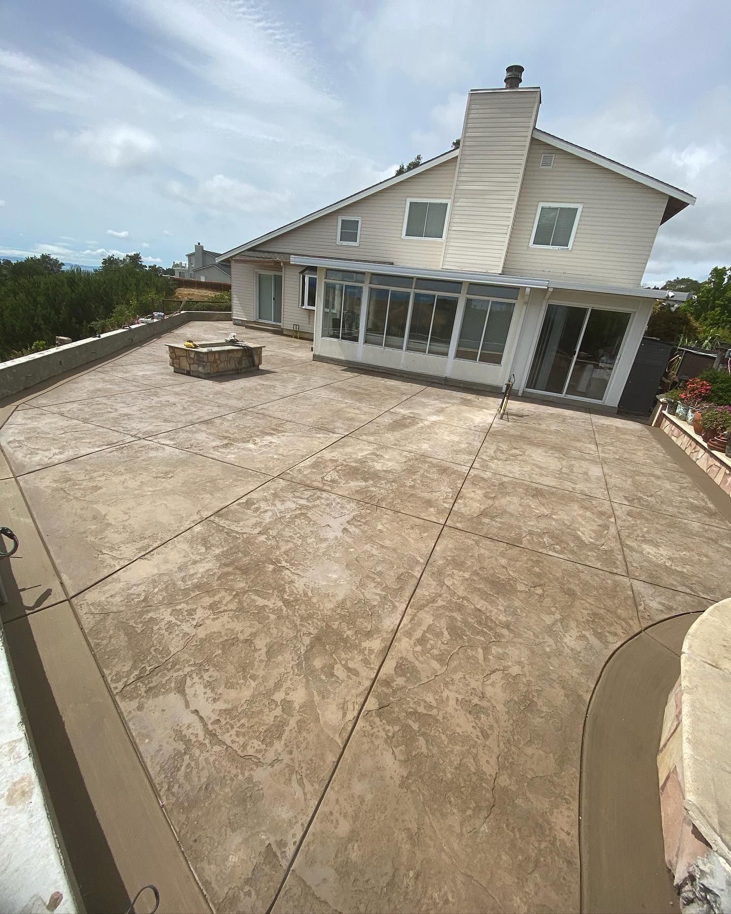 A tan, stamped-concrete patio with a square fire pit sits outside a two-story beige house with a sunroom.