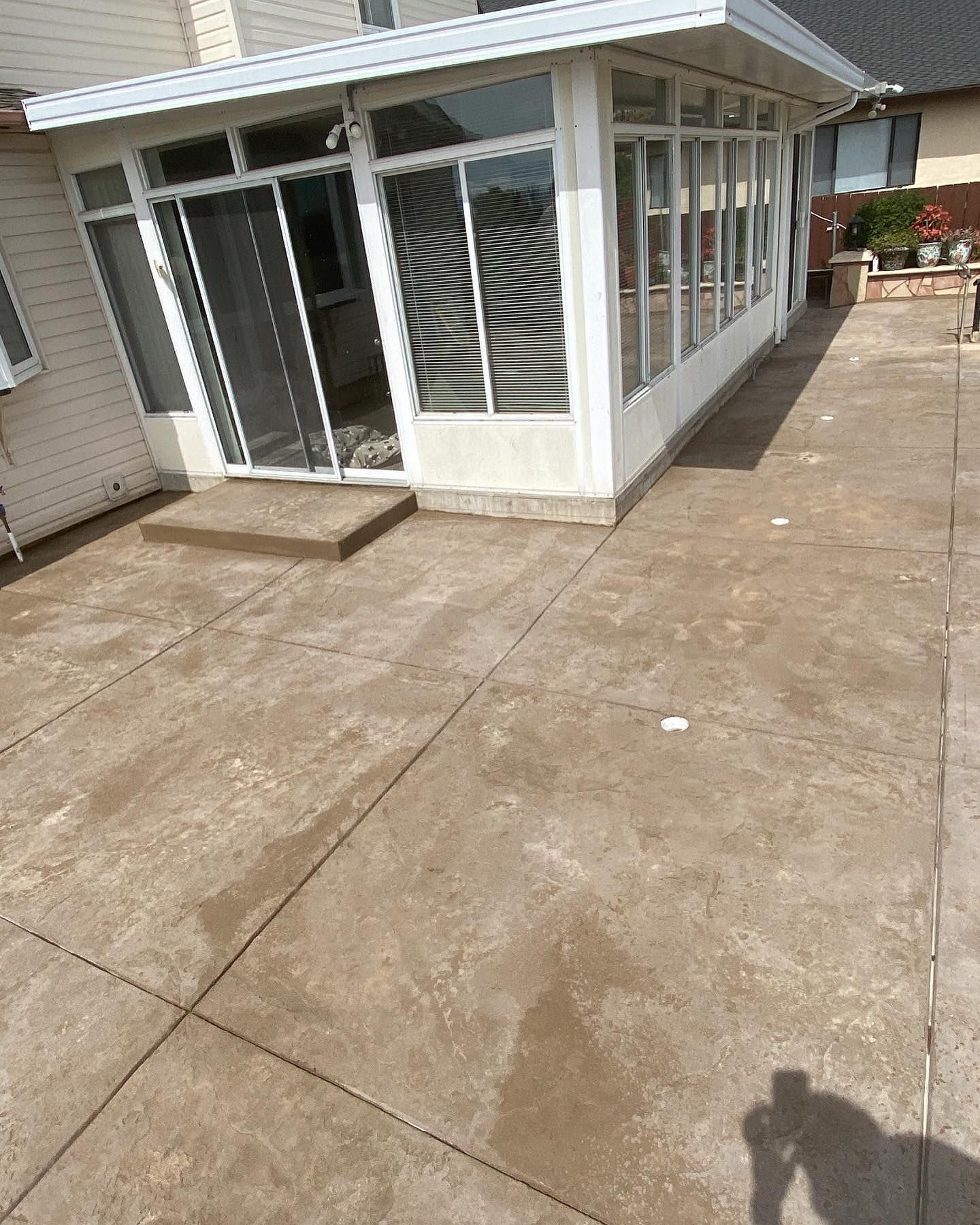 A sunroom with white frames and sliding glass doors sits on a large, rectangular concrete patio attached to a house.