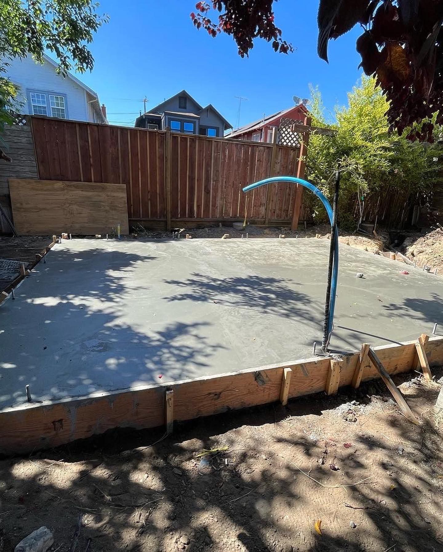 A fresh rectangular concrete slab with wooden formwork in a backyard, featuring a curved blue conduit pipe.