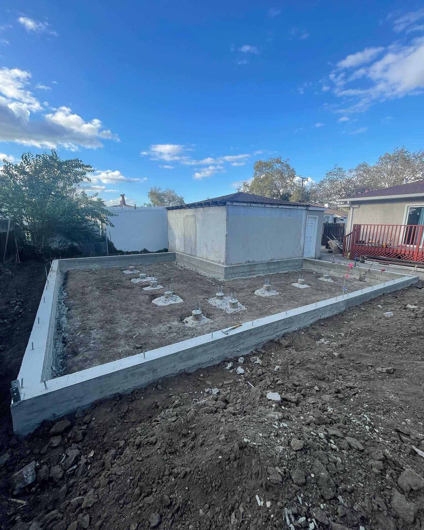 A concrete foundation frame for a new structure sits in a dirt yard next to an existing building under a clear blue sky.