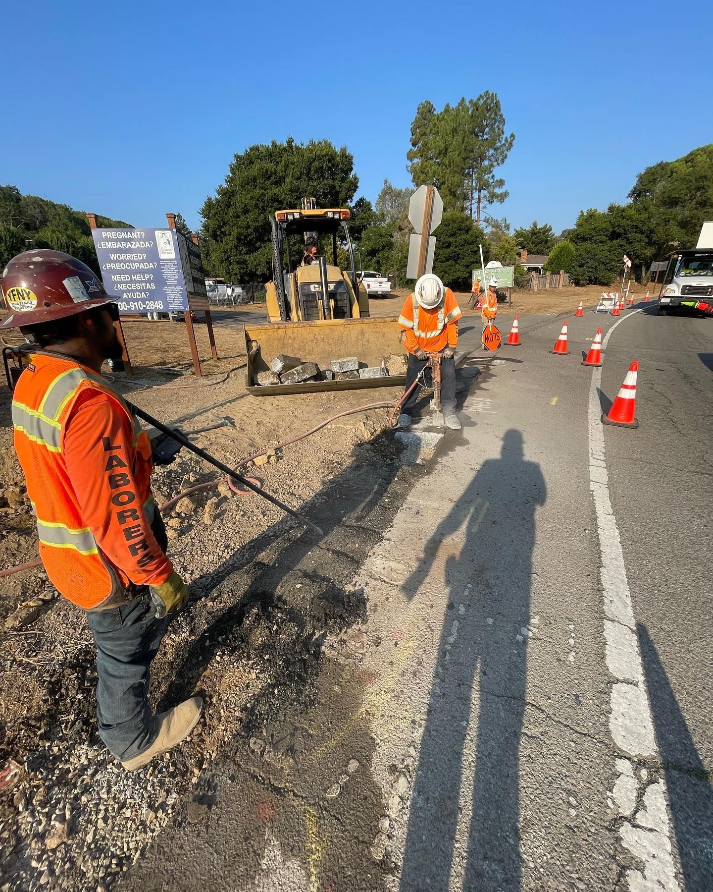 Two construction workers in high-visibility orange shirts work near a road with a bulldozer and traffic cones.