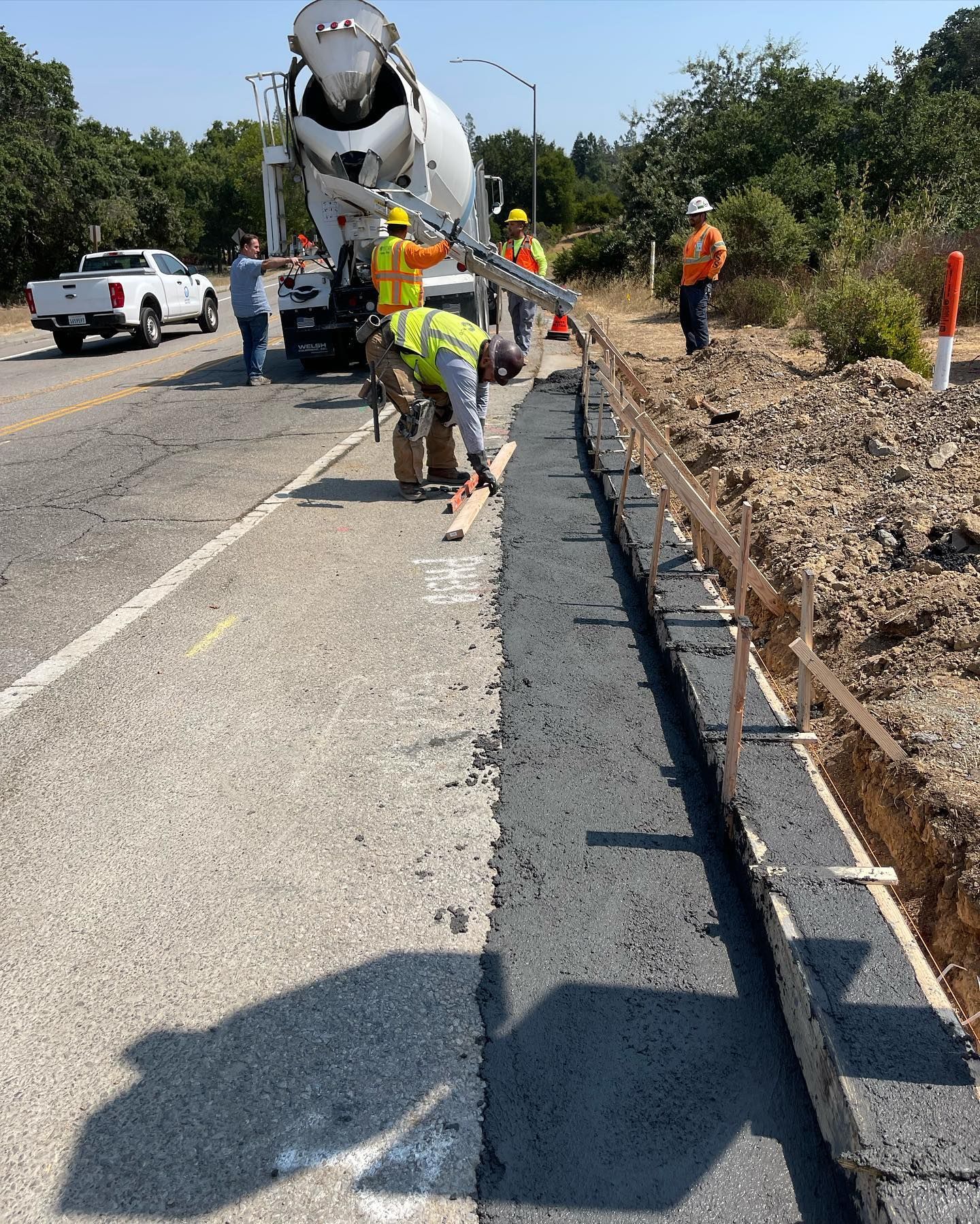 Construction workers use a concrete truck to pour a new curb along the edge of a paved road.