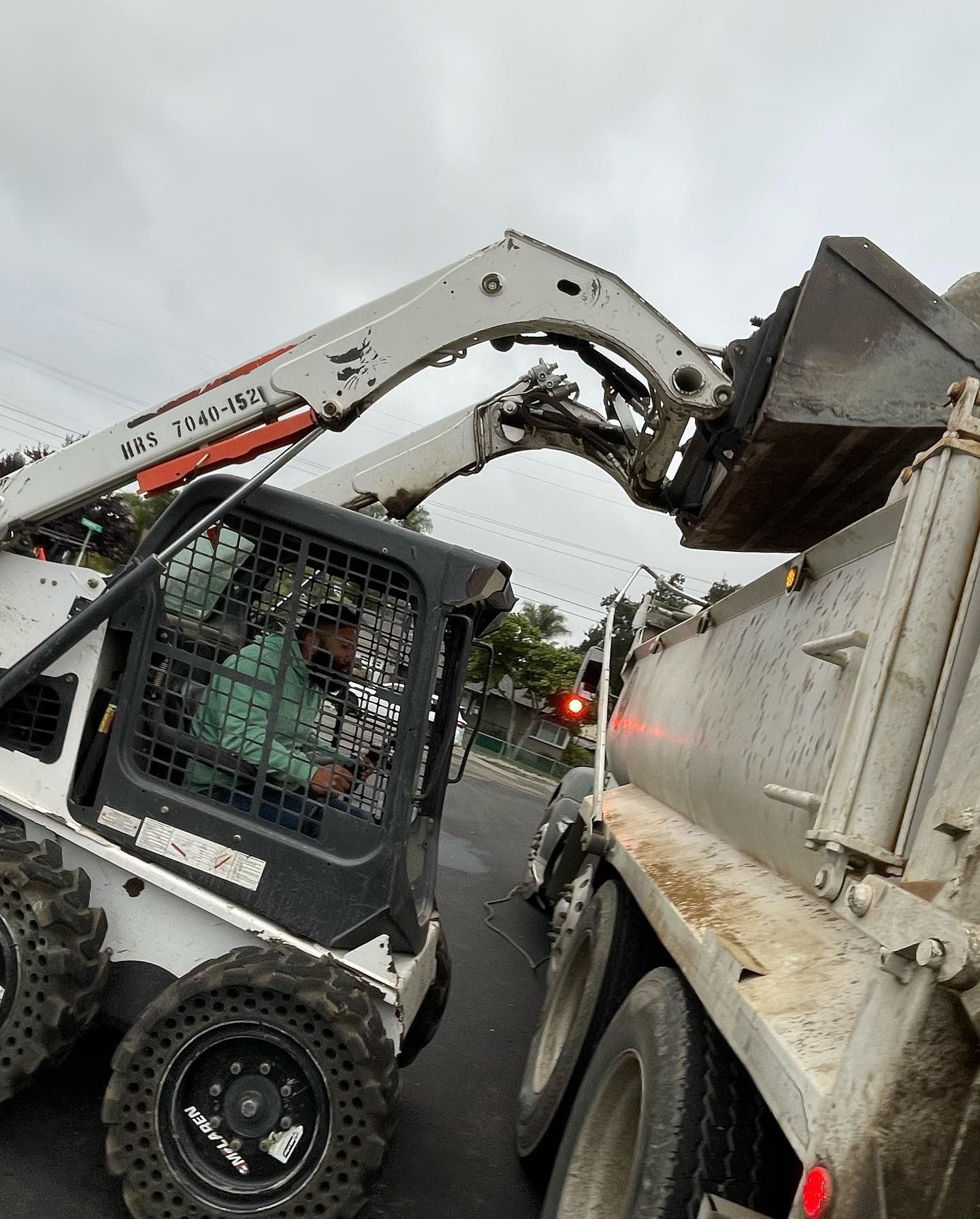A white skid steer loader lifts its bucket to dump material into the bed of a large dump truck on an overcast day.
