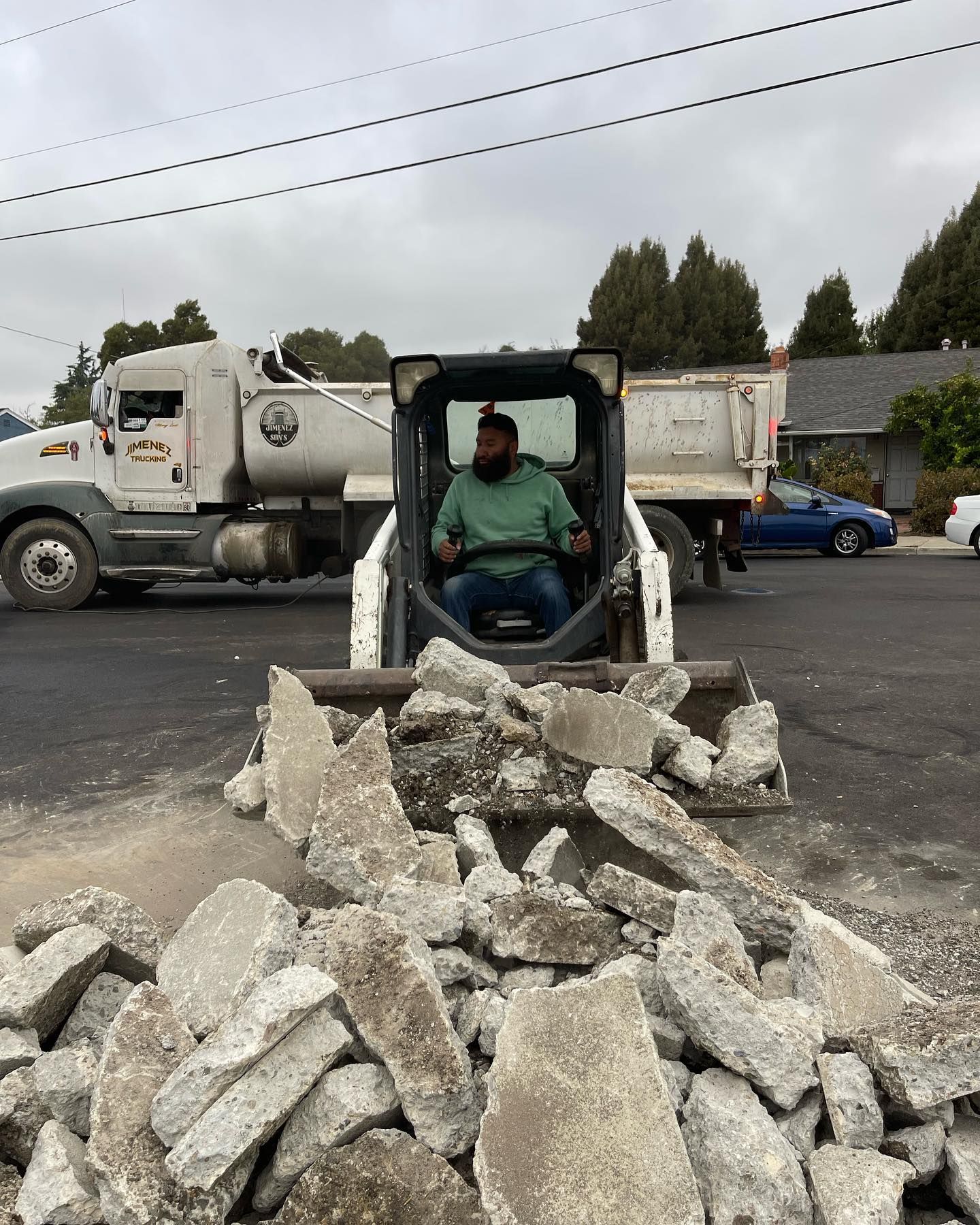 A person in a light green shirt operates a skid steer loader, moving a large pile of concrete rubble on an asphalt lot.