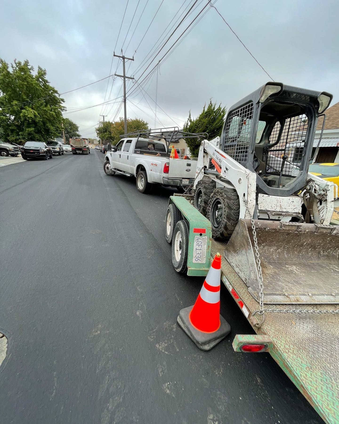 A white pickup truck towing a flatbed trailer with a skid steer parked on a newly paved road next to an orange traffic cone.