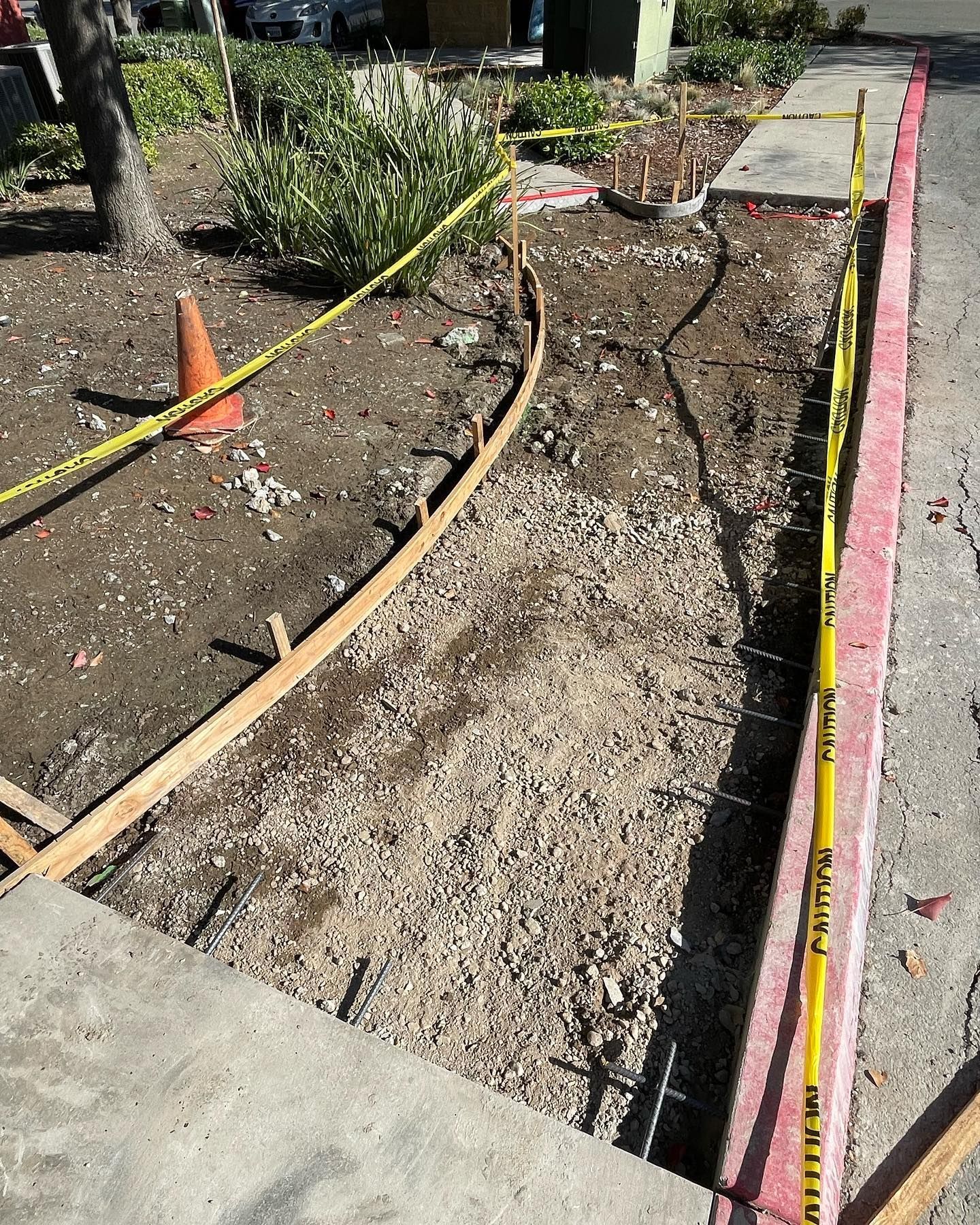 A construction site with wooden concrete forms, gravel ground, an orange cone, and yellow caution tape on a curb.