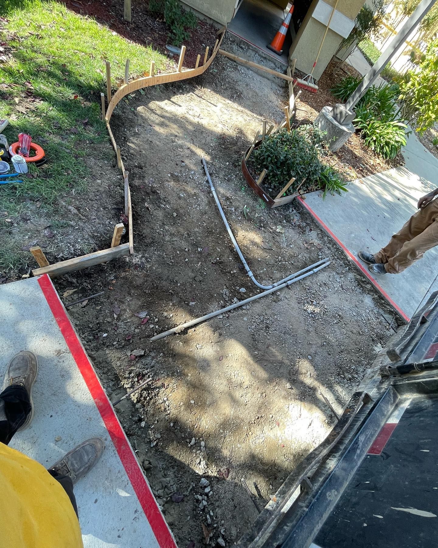Construction crew prepares a curved walkway area with wooden concrete forms, metal framing, and red safety tape.