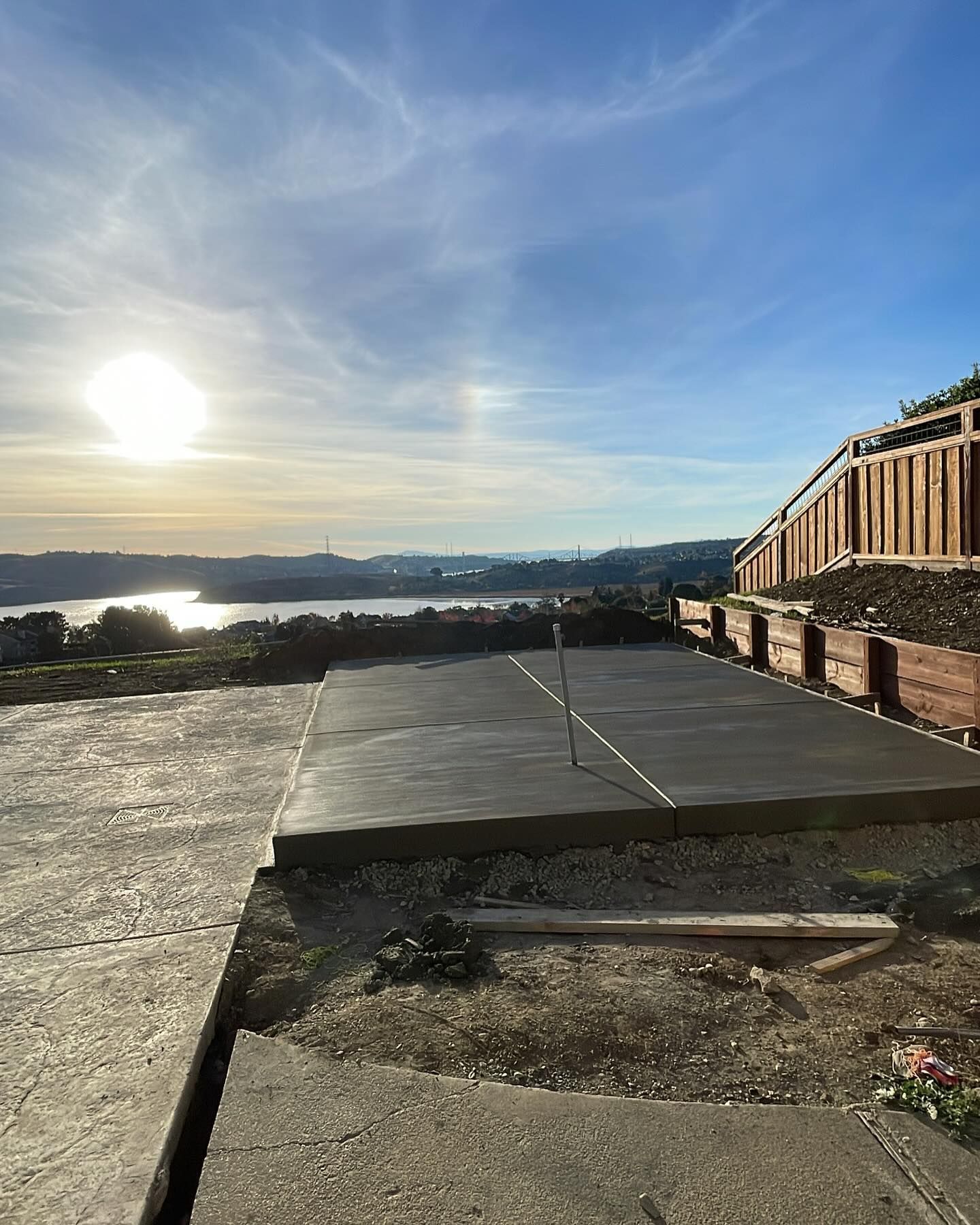 A concrete slab sits on a hillside overlooking a body of water at sunset, with a wooden fence along the right edge.