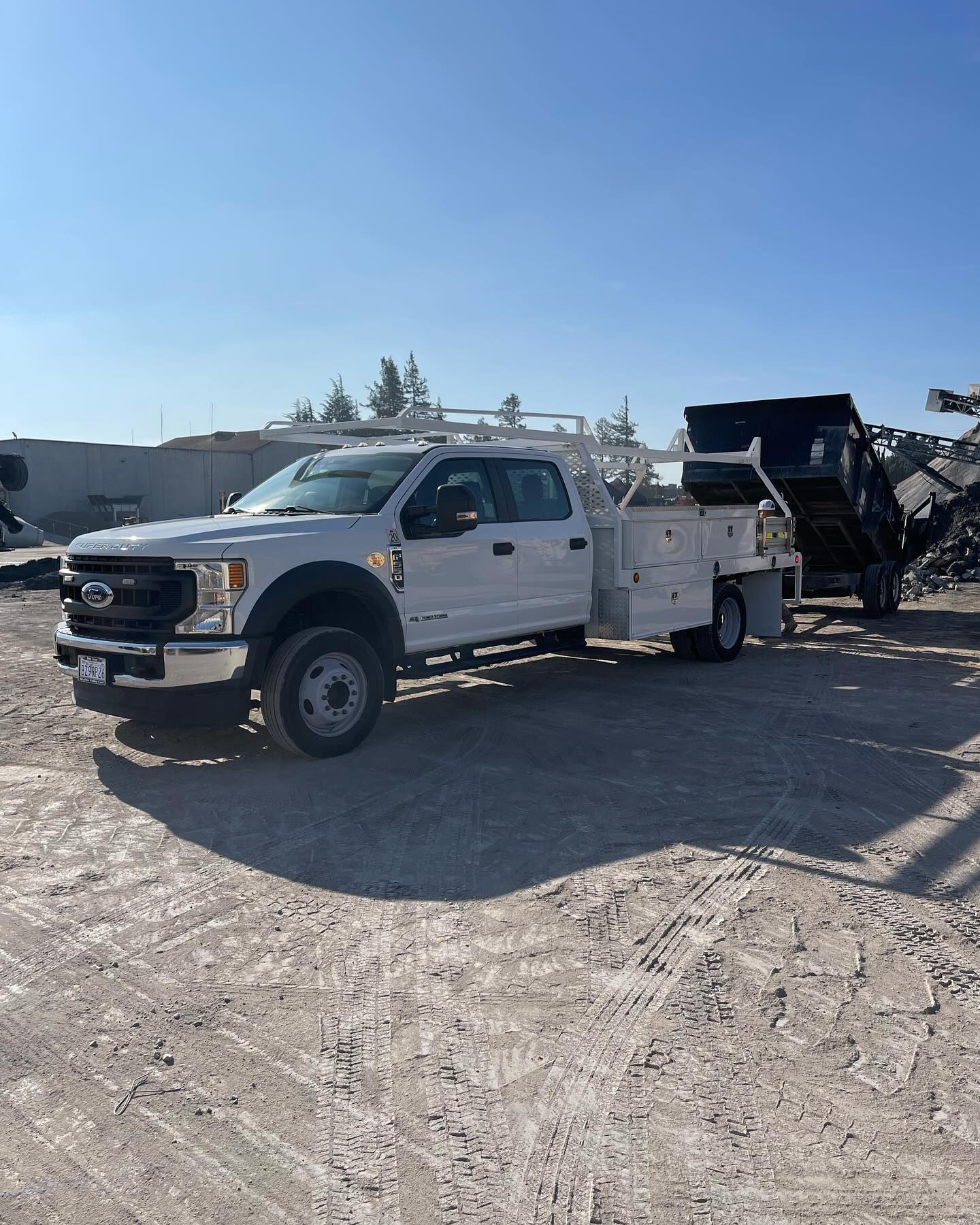 A white Ford Super Duty utility truck with a service body and overhead rack parked on a dirt lot, hitched to a trailer.