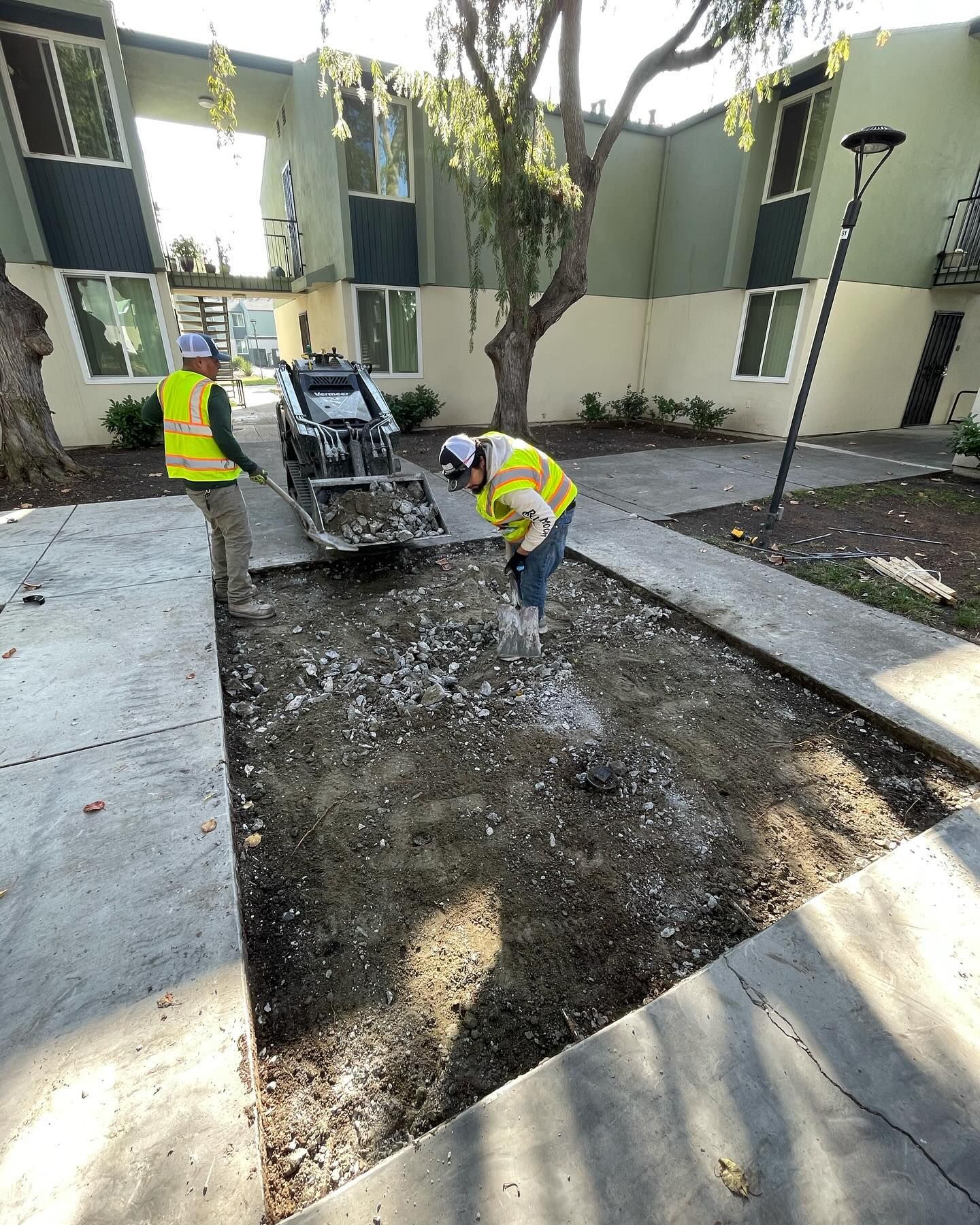 Two workers in high-visibility vests use a skid steer and hand tools to remove concrete between apartment buildings.